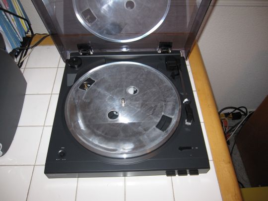 Photo shows a nearly-assembled Audio-Technica AT-LP2D-USB turntable. Prominent is a bare metal platter with four holes. The pulley that spins the platter is visible through one hole. The drive belt is over the pulley.