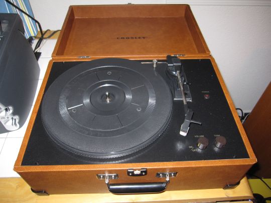 Photo shows the Crosley CR249 turntable on a table from above. The cover is open, revealing the platter and controls.