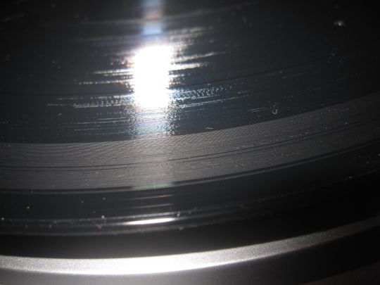 Close-up photo shows a black record with a gray ring starting from the outer edge and continuing through the first track. Damage was from a defective stylus on a Grace Digital Audio Vinylwriter (AVPUSB01S) USB turntable.